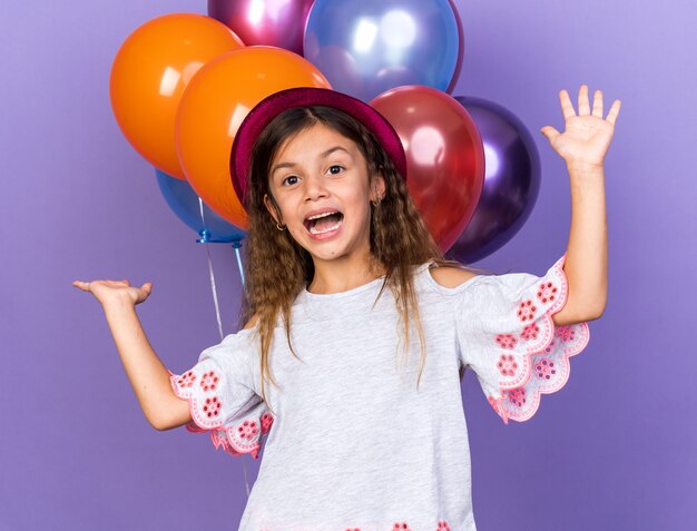 gioiosa piccola ragazza caucasica con cappello da festa viola in piedi con le mani alzate davanti a palloncini di elio isolati sul muro viola con spazio di copia