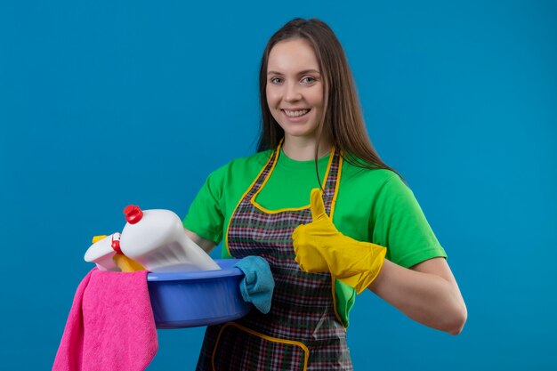 Gioiosa pulizia giovane donna che indossa l'uniforme in guanti che tengono gli strumenti di pulizia sulla parete blu isolata