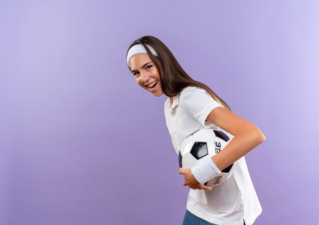 Gioiosa ragazza abbastanza sportiva che indossa fascia e braccialetto che tiene il pallone da calcio in piedi nella vista di profilo isolata sulla parete viola con spazio per le copie