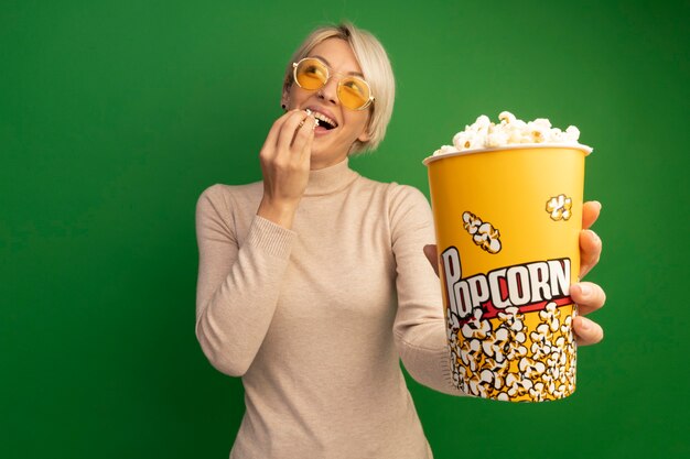 Gioiosa ragazza bionda che indossa occhiali da sole che allunga il secchio di popcorn e pezzi di popcorn vicino alla bocca guardando il lato isolato sulla parete verde con spazio di copia