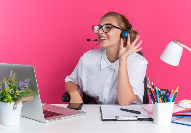 Gioiosa ragazza bionda del call center che indossa auricolare e occhiali seduto alla scrivania con strumenti di lavoro guardando la fotocamera che afferra l'auricolare isolato sul muro rosa