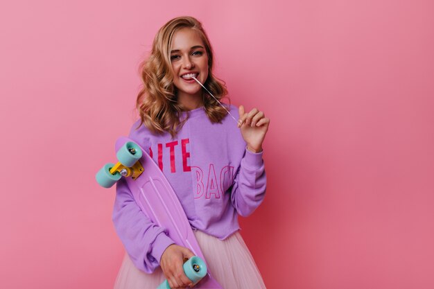 Gioiosa ragazza caucasica con gomma da masticare divertendosi. Incredibile donna bianca con lo skateboard in piedi sul rosa.