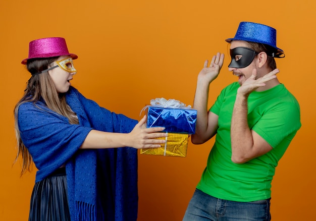 Gioiosa ragazza con cappello rosa che indossa una maschera per gli occhi in maschera con scatole regalo guardando un bell'uomo sorpreso con cappello blu che indossa una maschera per gli occhi in maschera alzando le mani guardando le scatole