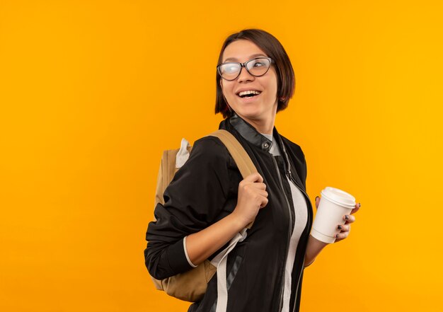 Gioiosa ragazza giovane studente con gli occhiali e borsa posteriore in piedi in vista di profilo guardando dietro tenendo la tazza di caffè isolata sull'arancio