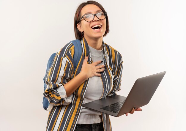 Gioiosa ragazza giovane studente con gli occhiali e borsa posteriore tenendo il computer portatile con la mano sul petto isolato su bianco
