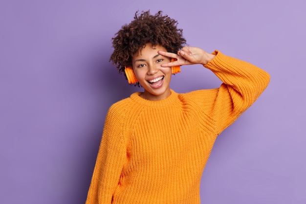 Gioiosa ragazza millenaria dalla pelle scura rilassata fa il gesto di vittoria ascolta la musica preferita in cuffie wireless indossa un maglione arancione lavorato a maglia