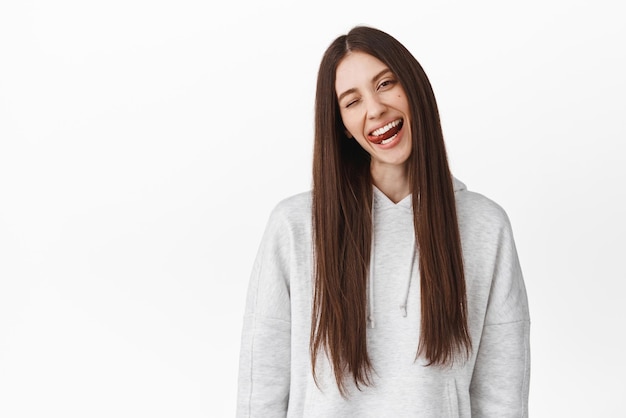 Gioiosa studentessa adolescente con lunghi capelli castani inclina la testa strizzando l'occhio a te e mostra la lingua positiva esprimono felicità e gioia in piedi su sfondo bianco