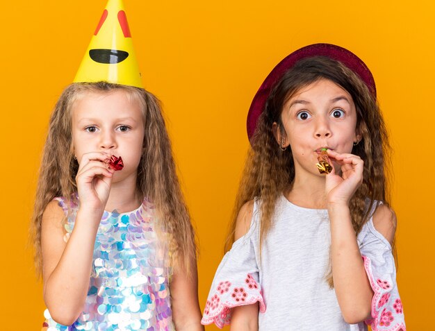 gioiose bambine graziose con cappelli da festa che soffia fischietti isolati sulla parete arancione con spazio di copia