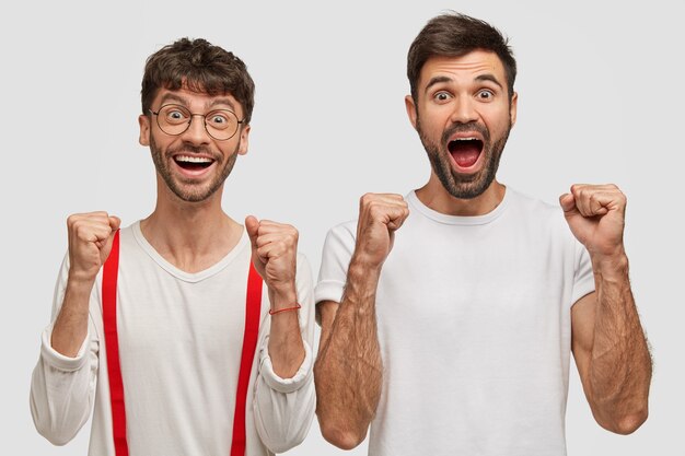 Gioiosi con la barba lunga due giovani stringono i pugni e gridano di felicità, vestiti casualmente, isolati sul muro bianco