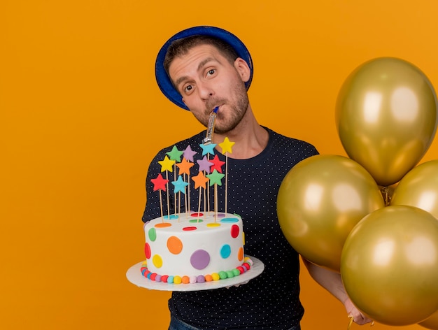 Gioioso bell'uomo caucasico che indossa il cappello blu del partito tiene palloncini di elio e torta di compleanno che soffia fischio isolato su sfondo arancione con spazio di copia