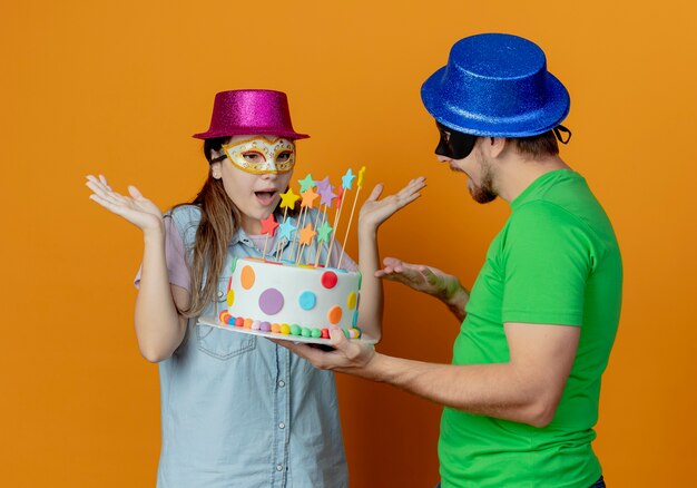 Gioioso bell'uomo con cappello blu che indossa una maschera per gli occhi in maschera con una torta di compleanno che guarda una ragazza sorpresa che indossa un cappello rosa e una maschera per gli occhi in maschera alzando le mani guardando la torta