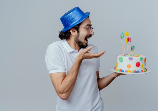 Gioioso bell'uomo con gli occhiali e cappello blu che tiene e punti con la mano alla torta isolato su priorità bassa bianca