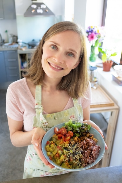 Gioioso blogger alimentare che presenta piatto di verdure fatto in casa, in piedi in cucina, che guarda l'obbiettivo e sorridente. Colpo verticale, angolo alto. Concetto di mangiare sano