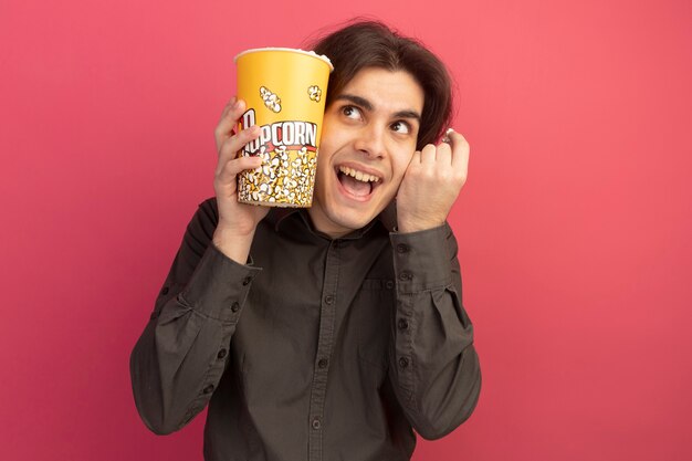 Gioioso che osserva in su giovane bel ragazzo che indossa la maglietta nera che tiene secchio di popcorn con pace popcorn isolato sulla parete rosa