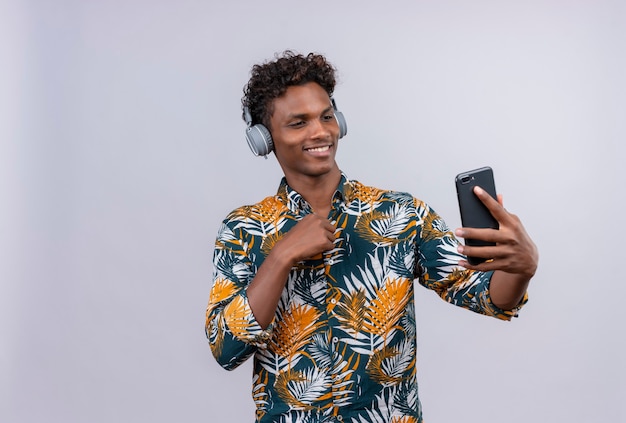 Gioioso e sorridente giovane bello di carnagione scura con capelli ricci in foglie camicia stampata che indossa le cuffie guardando il suo telefono cellulare su uno sfondo bianco