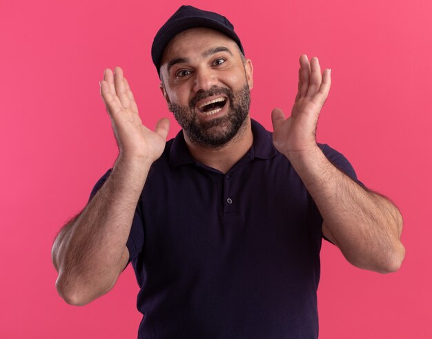 Gioioso fattorino di mezza età in uniforme e berretto che si tiene per mano intorno al viso isolato sul muro rosa