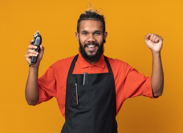 Gioioso giovane barbiere afroamericano che indossa l'uniforme che tiene i tagliacapelli facendo un gesto forte isolato sulla parete arancione