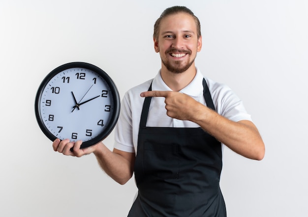 Gioioso giovane barbiere bello che indossa la tenuta uniforme e indicando l'orologio isolato su bianco