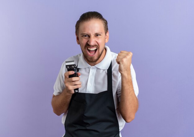 Gioioso giovane barbiere bello che indossa uniforme che tiene i tagliacapelli e il pugno di serraggio isolato sulla porpora