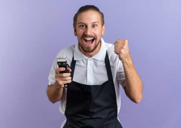 Gioioso giovane barbiere bello che indossa uniforme che tiene i tagliacapelli e il pugno di serraggio isolato sulla porpora