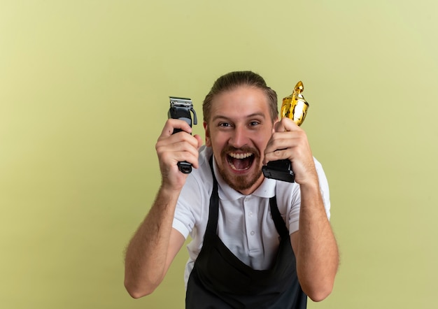 Gioioso giovane barbiere bello che tiene tagliacapelli e tazza del vincitore isolato su verde oliva