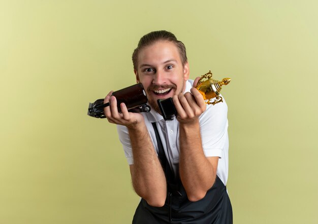 Gioioso giovane barbiere bello che tiene tagliacapelli, flacone spray e tazza del vincitore isolato su verde oliva