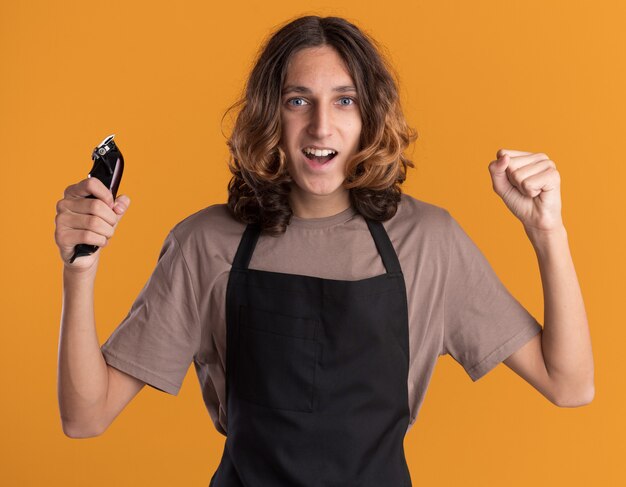 Gioioso giovane bel barbiere che indossa l'uniforme che tiene i tagliacapelli guardando davanti facendo sì gesto isolato sulla parete arancione