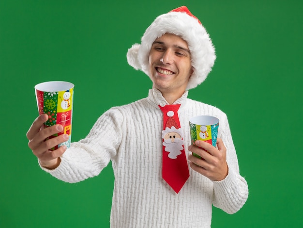 Gioioso giovane bel ragazzo che indossa cappello di natale e cravatta di babbo natale che tiene e allunga le tazze di plastica di natale guardando uno di loro isolato sulla parete verde