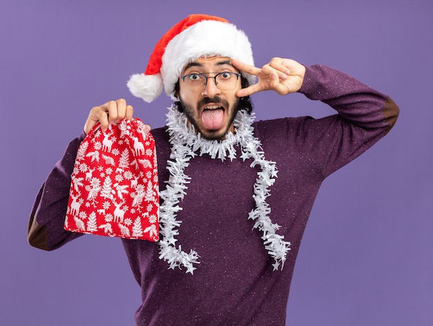 Gioioso giovane bel ragazzo che indossa il cappello di Natale con la ghirlanda sul collo che tiene la borsa di Natale che mostra la lingua e il gesto di pace isolato sulla parete blu