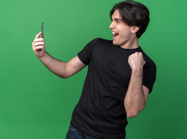 Gioioso giovane bel ragazzo che indossa una t-shirt nera che tiene in mano e guarda il telefono mostrando sì gesto isolato sul muro verde