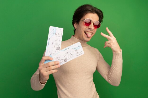 Gioioso giovane bel ragazzo con gli occhiali che tengono i biglietti che mostrano sì gesto isolato sulla parete verde