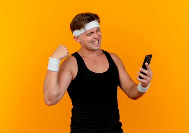 Gioioso giovane bell'uomo sportivo che indossa la fascia e braccialetti che tengono e guardando il telefono cellulare e gesticolando forte isolato sull'arancio