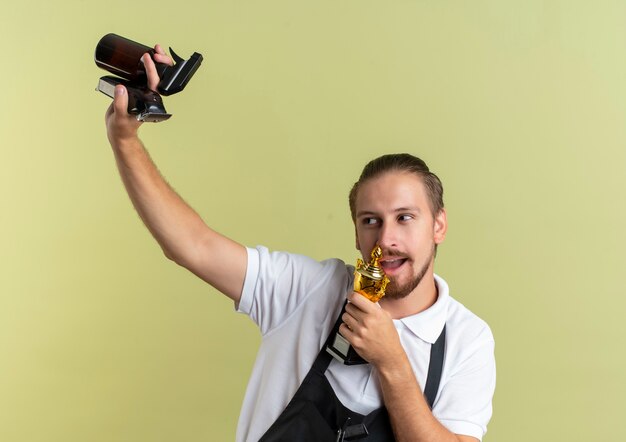 Gioioso giovane bello barbiere alzando il tagliacapelli, flacone spray e fingere di cantare utilizzando la coppa del vincitore come microfono guardando il lato isolato su verde oliva