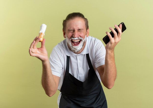 Gioioso giovane bello barbiere che indossa uniforme tenendo il telefono cellulare e pennello da barba con gli occhi chiusi e con crema da barba applicata sulla sua barba isolata su verde oliva