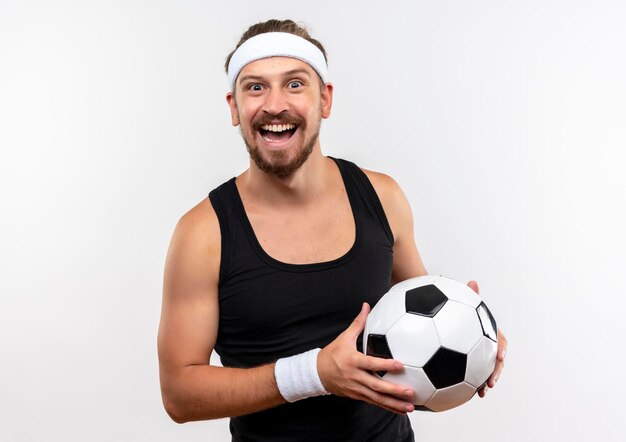 Gioioso giovane bello sportivo che indossa fascia e braccialetti che tengono un pallone da calcio che sembra isolato su un muro bianco