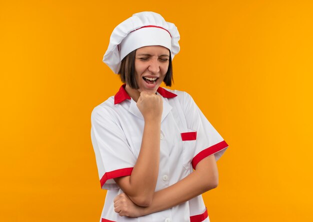 Gioioso giovane cuoco femminile in uniforme del cuoco unico che mette il pugno sotto il mento con gli occhi chiusi isolati sull'arancio