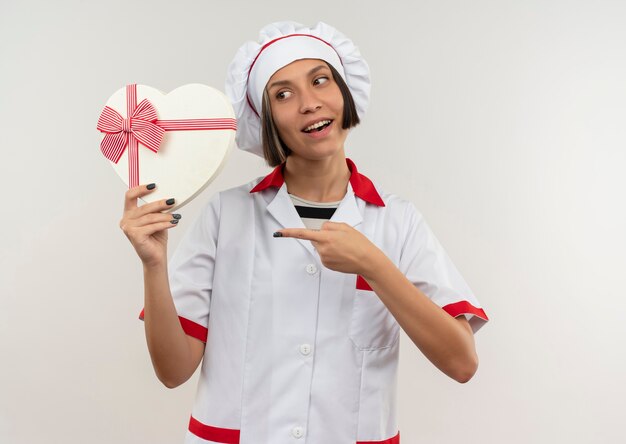 Gioioso giovane cuoco femminile in uniforme del cuoco unico che tiene guardando e indicando il contenitore di regalo a forma di cuore isolato su bianco