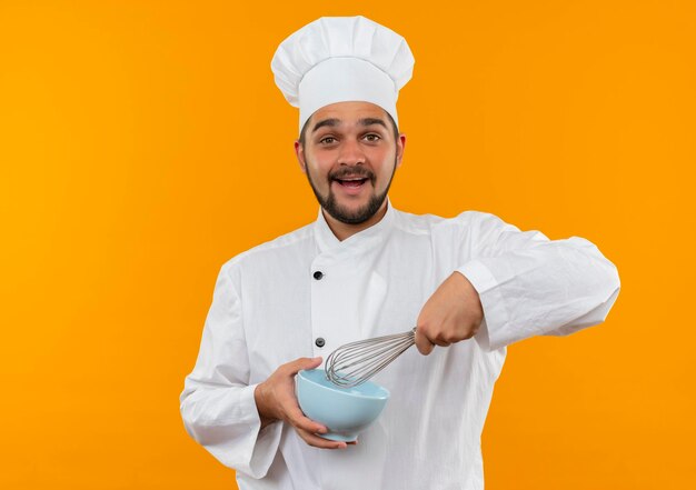 Gioioso giovane cuoco maschio in uniforme da chef che tiene in mano una frusta e una ciotola isolate sulla parete arancione con spazio per le copie