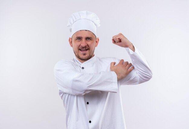 Gioioso giovane e bello cuoco in uniforme da chef che gesticola forte e mette la mano sulla spalla isolata sul muro bianco