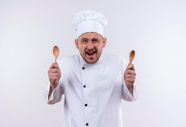 Gioioso giovane e bello cuoco in uniforme da chef che tiene in mano i cucchiai sul muro bianco isolato