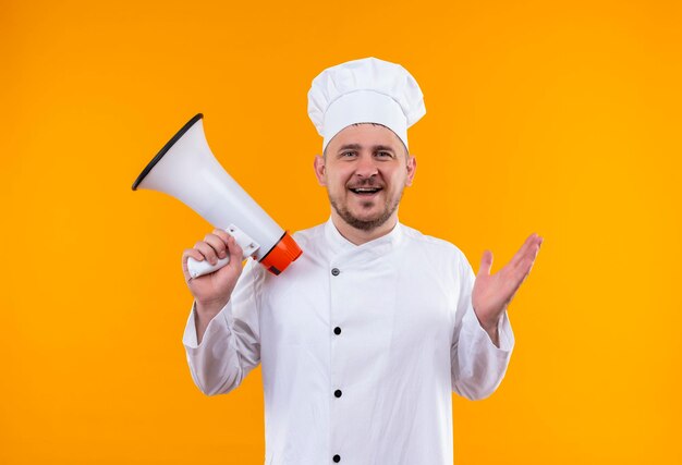 Gioioso giovane e bello cuoco in uniforme da chef che tiene in mano un altoparlante isolato su una parete arancione