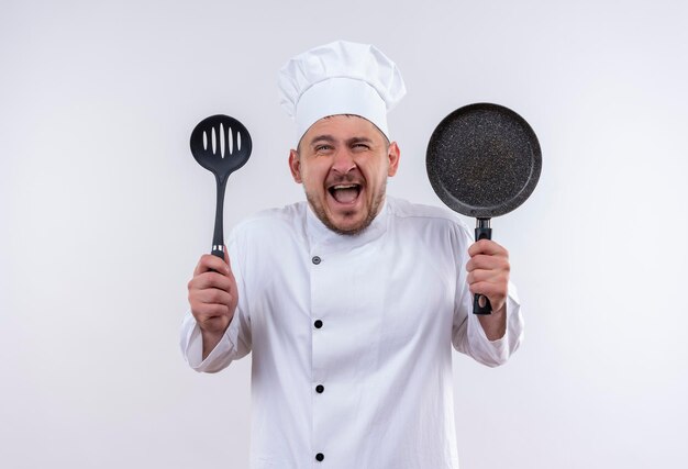 Gioioso giovane e bello cuoco in uniforme da chef che tiene in mano un cucchiaio scanalato e una padella sul muro bianco isolato