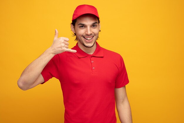 gioioso giovane fattorino che indossa uniforme e cappuccio guardando la fotocamera che mostra il gesto di chiamata isolato su sfondo giallo