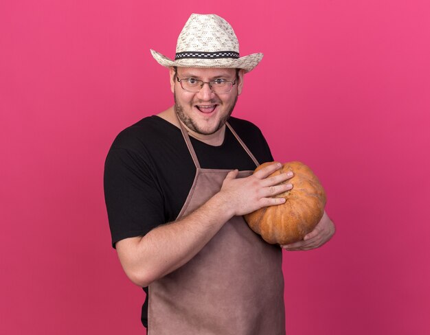 Gioioso giovane giardiniere maschio che indossa il cappello da giardinaggio tenendo la zucca isolata sulla parete rosa