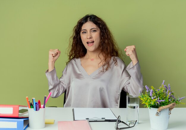 Gioioso giovane impiegato abbastanza femminile che si siede allo scrittorio con gli strumenti dell'ufficio che mostrano il gesto di sì isolato su fondo verde oliva