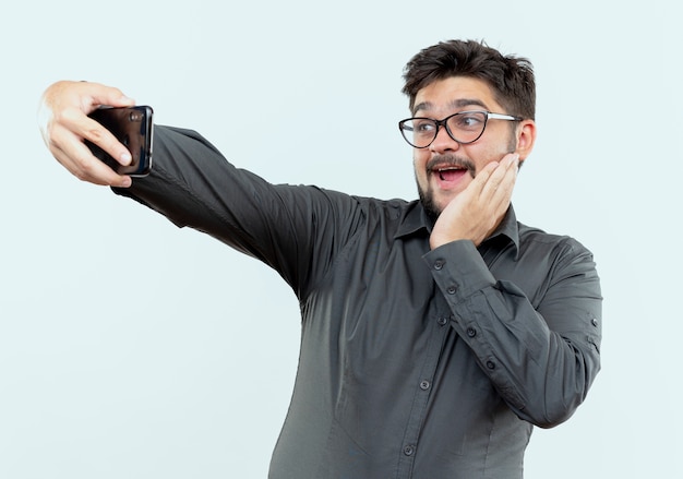 Gioioso giovane imprenditore con gli occhiali prendere un selfie e mettendo la mano sulla guancia isolati su sfondo bianco