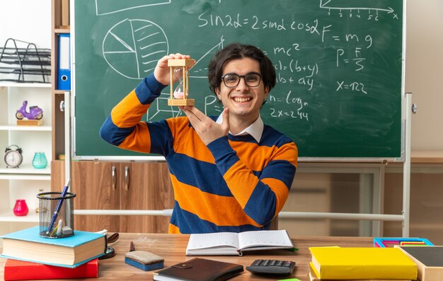 Gioioso giovane insegnante di geometria caucasica con gli occhiali seduto alla scrivania con strumenti scolastici in aula con clessidra