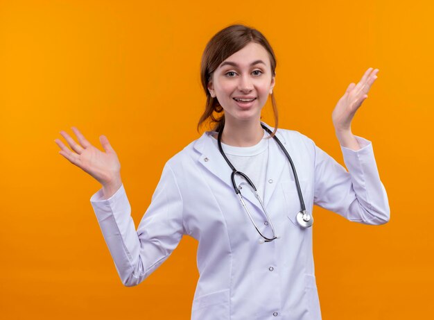 Gioioso giovane medico femminile che indossa abito medico e stetoscopio con le braccia aperte sullo spazio arancione isolato