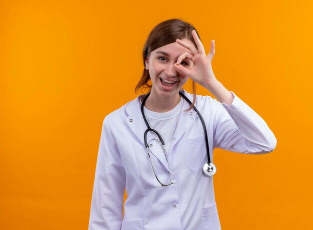 Gioioso giovane medico femminile che indossa abito medico e stetoscopio facendo gesto di sguardo sullo spazio arancione isolato con spazio di copia