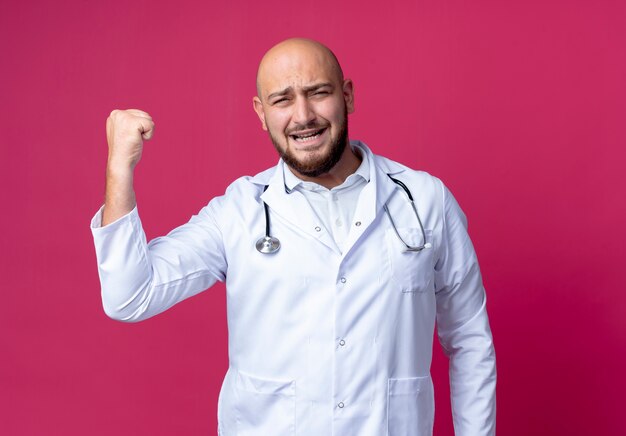 Gioioso giovane medico maschio calvo che indossa veste medica e stetoscopio che mostra un forte gesto isolato sulla parete rosa